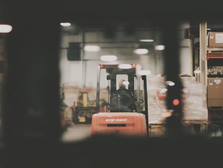 Unscharfe Aufnahme eines Lagerhauses, im Fokus ein Gabelstapler mit einem Fahrer, der Paletten mit Waren transportiert. Umgeben von Regalen mit Kartons und verpackten Produkten. Die Szene wirkt geschäftig und industriell.