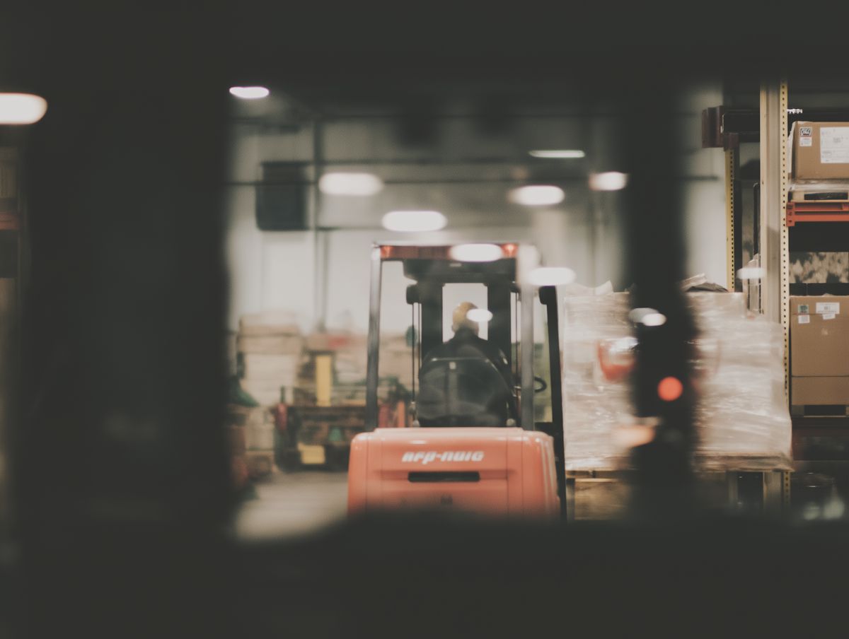 Unscharfe Aufnahme eines Lagerhauses, im Fokus ein Gabelstapler mit einem Fahrer, der Paletten mit Waren transportiert. Umgeben von Regalen mit Kartons und verpackten Produkten. Die Szene wirkt geschäftig und industriell.