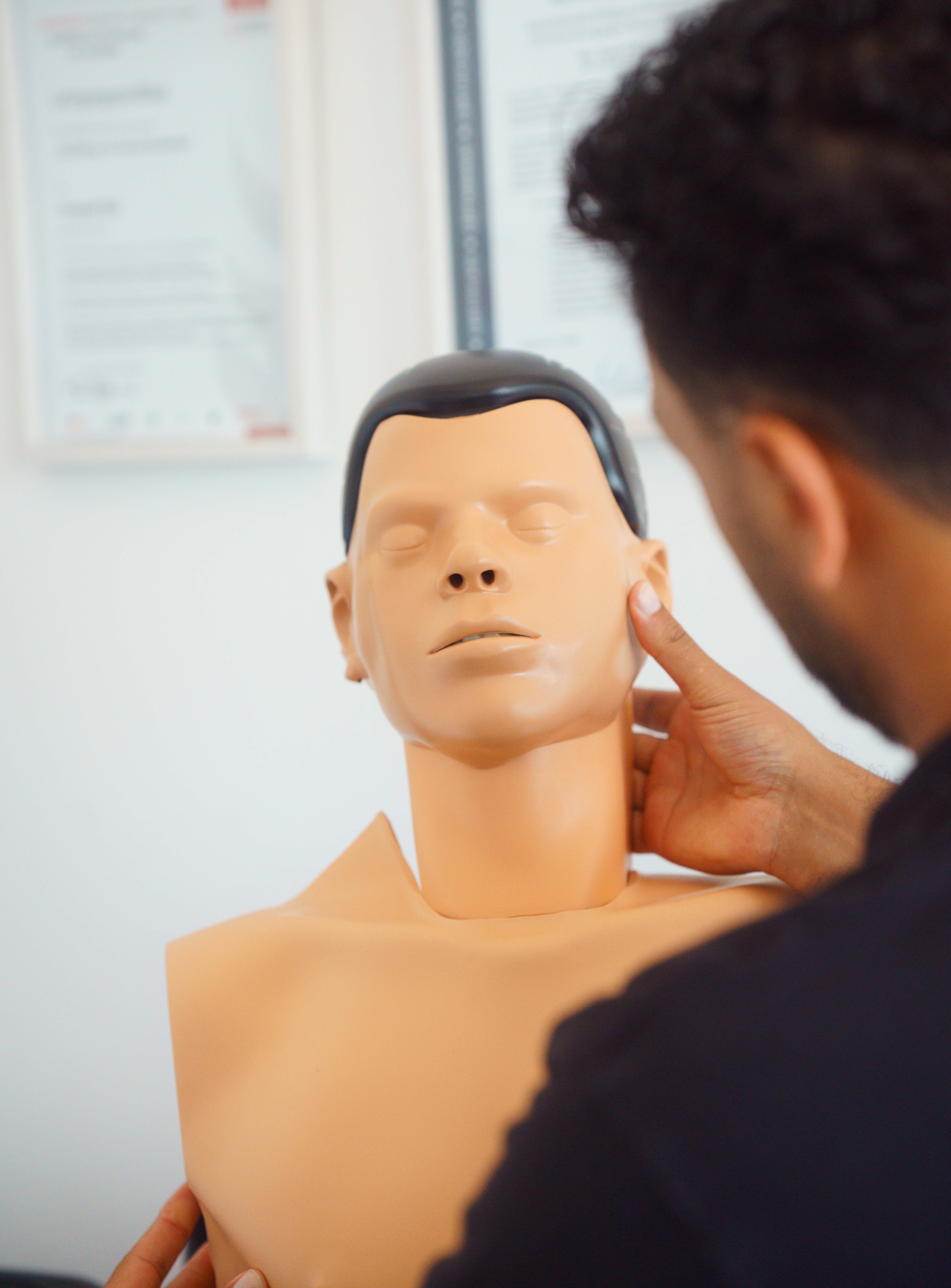 Ein Mann demonstriert an einer Erste-Hilfe-Übungspuppe das Überprüfen der Atmung, indem er den Kopf der Puppe überstreckt und das Kinn anhebt. Die Szene findet in einem Schulungsraum statt, im Hintergrund sind unscharfe Zertifikate oder Infotafeln an der Wand zu erkennen.